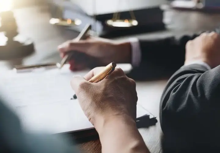Dłonie piszące długopisami na dokumentach, w tle waga sprawiedliwości.