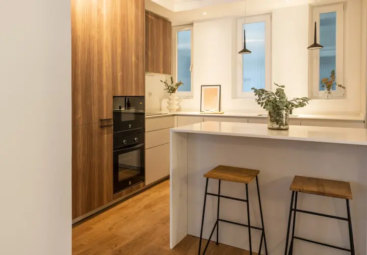 Cocina moderna con muebles de madera, encimera blanca y dos taburetes. La luz fría k ilumina el espacio, creando un ambiente acogedor.