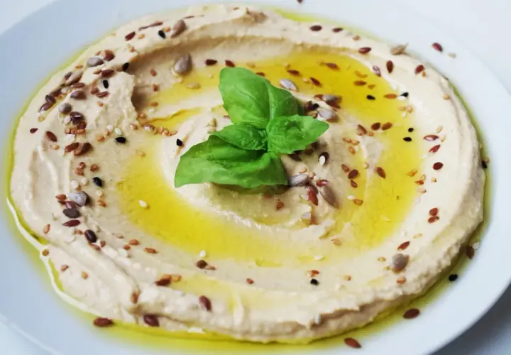 Hummus di ceci cremoso, condito con olio d'oliva, semi misti e una foglia di basilico. Perfetto da gustare con pane pita o verdure fresche.