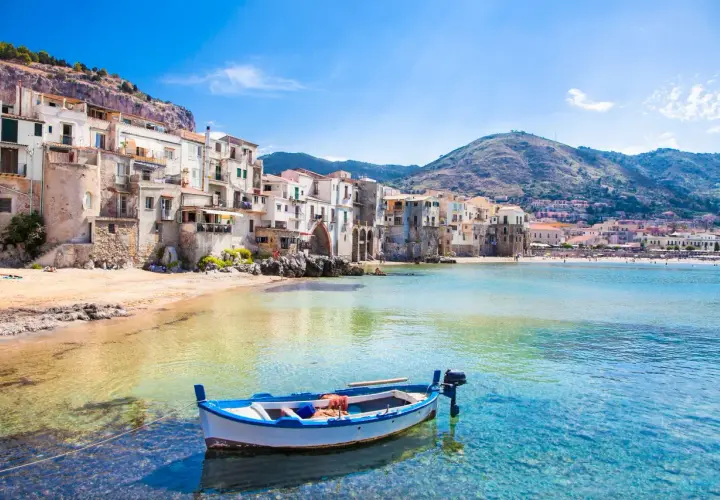 A charming fishing boat bobs in clear turquoise waters off the coast of Cefalù, a picturesque town with colorful buildings cascading down a hillside, a must-see among places to visit in Sicily.