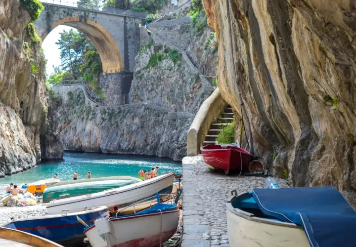 Boats line the shore of Fiordo di Furore, a stunning fjord with turquoise waters and a dramatic arched bridge overhead.