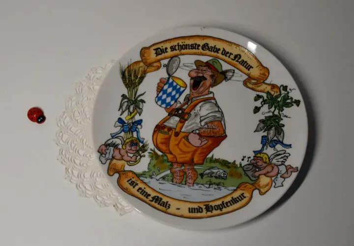 Ein Teller mit einem fröhlichen Mann im Lederhosen, der ein Bier hält. Die Inschrift lautet: "Die schönste Gabe der Natur ist eine Malz- und Hopfenkur." Judith Merkle Riley hat dieses Motiv entworfen.