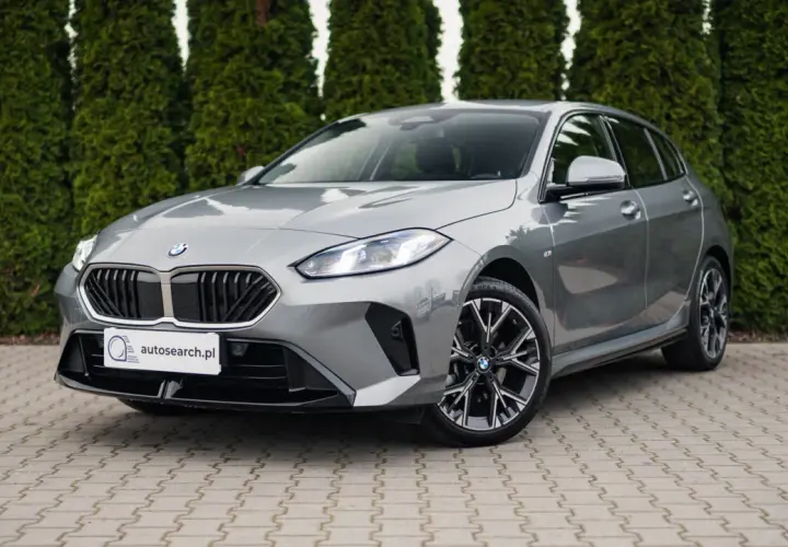 Szare BMW 118d z czarnym grillem i felgami stoi przed żywopłotem.
