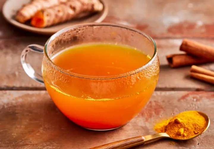 Ein Glas leuchtend orangefarbener Kurkuma-Tee steht auf einem Holztisch. Daneben liegen frische Kurkumawurzeln, Zimtstangen und ein Löffel mit Kurkumapulver. Die Wirkung von Kurkuma-Tee ist bekannt.