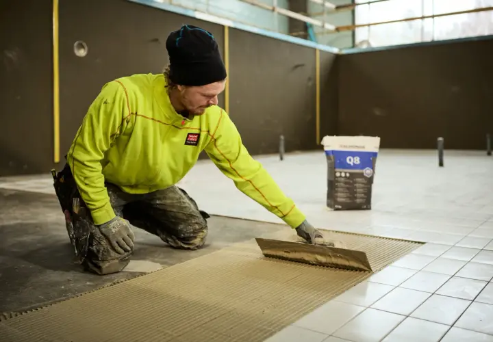 Man i gul jacka och mössa sprider ut fix med en tandspackel för att byta golv. En säck med fix står i bakgrunden.