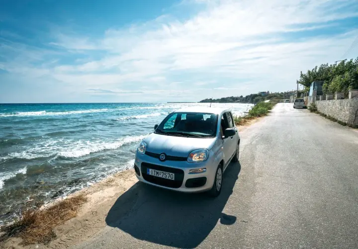 A silver Fiat Panda parked on a coastal road in Greece. The turquoise sea laps the shore beside the car, suggesting this is the perfect way to explore.