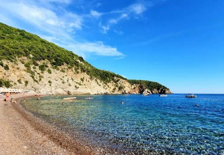 Kamienista plaża w Czarnogórze z turkusową wodą i zielonym wzgórzem w tle.