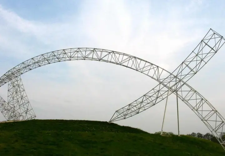Metalowa brama w kształcie ryby, wznosząca się na zielonym wzgórzu pod błękitnym niebem.