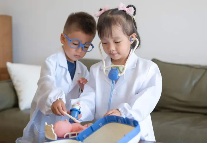 Dos niños, vestidos de doctores, atienden a un muñeco. La niña usa un estetoscopio, mientras el niño le administra una inyección. Es una escena de juego de roles, una brecha de Rolando con niños.