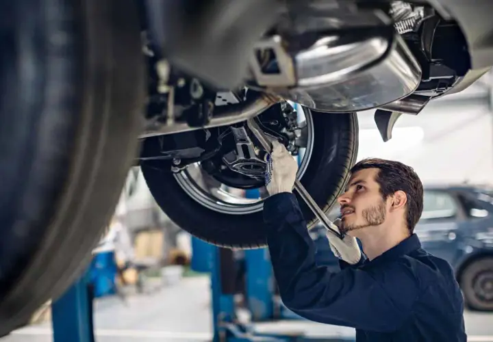 Meccanico con guanti bianchi lavora sotto un'auto sollevata, cercando la causa di un rumore metallico da sotto la macchina.