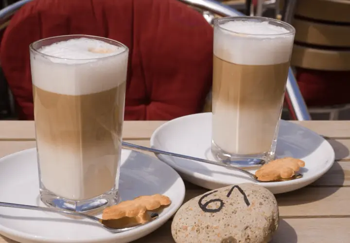 Dwa latte macchiato, czyli kawy z warstwami mleka i pianki, na stoliku. Obok ciasteczka i kamień z numerem 61.