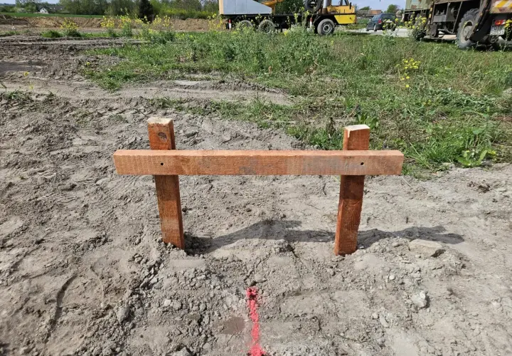 Drewniany palik z poprzeczką wyznacza przyszłą powierzchnię zabudowy. W tle widać plac budowy z maszynami.