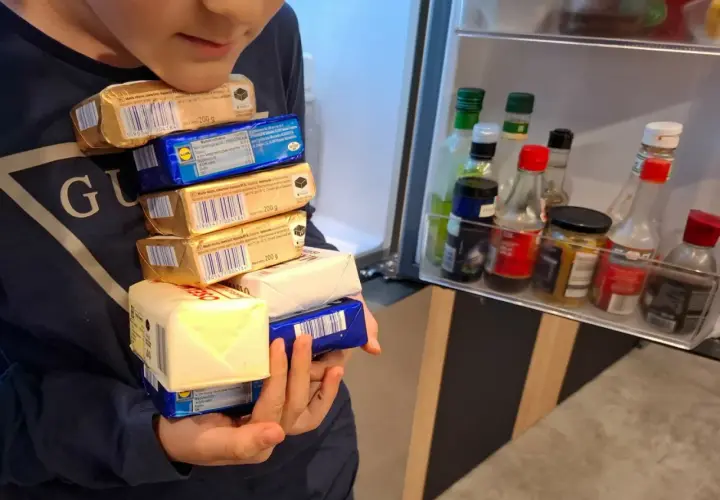 Dziecko trzyma stos opakowań masła przed otwartą lodówką z butelkami sosów.