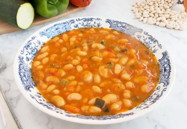 Un plato de alubias con verduras, cocinadas en una salsa rojiza y servidas en una vajilla con borde azul.
