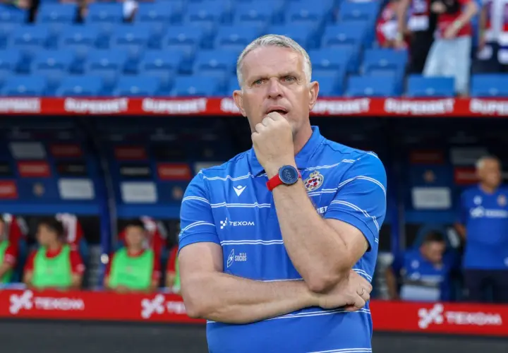 Trener Wisły Kraków w niebieskiej koszulce z białymi pasami, zamyślony, opiera dłoń na ustach.