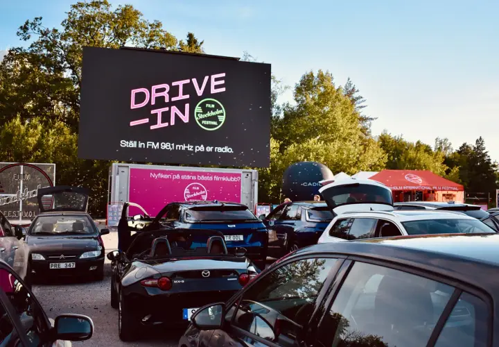Bilar samlade vid en solvalla drive in bio. Stor skärm visar "DRIVE-IN FILM Stockholm Festival".