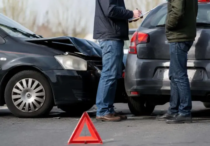 Dwaj mężczyźni spisują protokół po kolizji. Zniszczona maska czarnego auta i czerwony trójkąt ostrzegawczy sugerują, że czeka ich mandat za kolizje.