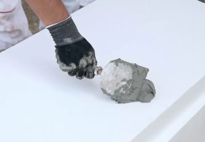 Ręka w rękawicy nakłada klej budowlany w piance na biały styropian.