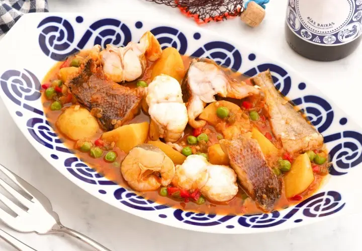 Caldeirada gallega servida en plato blanco con borde azul. Contiene trozos de pescado, patatas, guisantes y pimientos en una salsa rojiza.