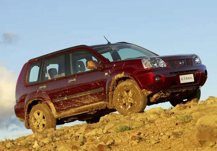 Czerwony Nissan X-Trail ubrudzony błotem podjeżdża pod skaliste wzniesienie.