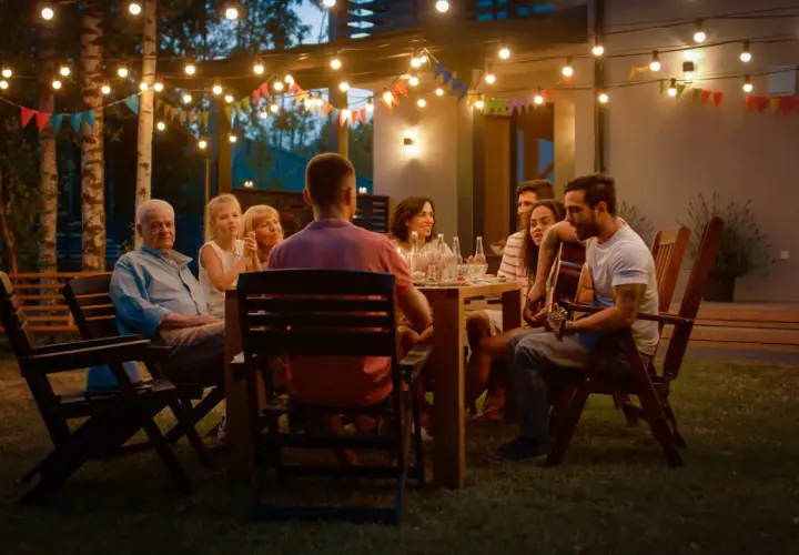 Rodzina i przyjaciele cieszą się wieczorem przy grillu. W tle girlandy światełek i kolorowe chorągiewki tworzą magiczną atmosferę. Idealna garden party stylizacja na grilla.