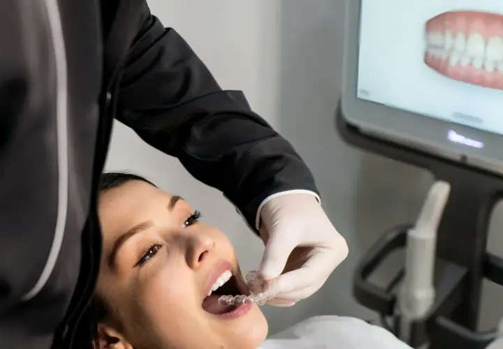 Dentysta w białej rękawiczce zakłada pacjentce przezroczyste nakładki. Na ekranie widoczny jest model uzębienia. Znieczulenie powierzchniowe nie jest widoczne, ale proces może być jego częścią.