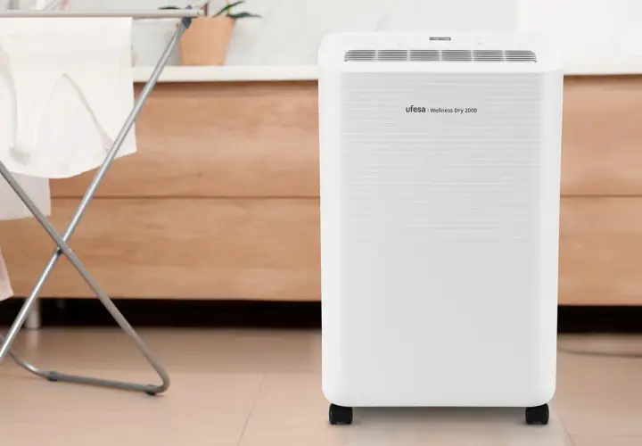 Un deshumidificador Ufesa Wellness Dry 2000, blanco y con ruedas, junto a un tendedero con ropa.
