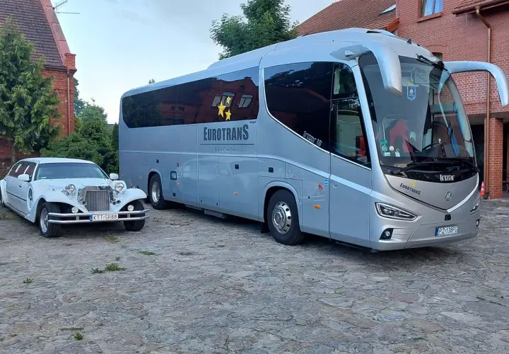 Biała limuzyna i szary autokar stoją obok siebie na brukowanym dziedzińcu.