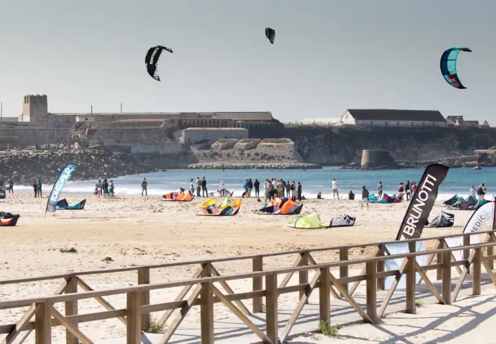 Plaża pełna kolorowych latawców i ludzi, gotowych do **kitesurfingu w Polsce**. W tle widać zabytkowe budowle.