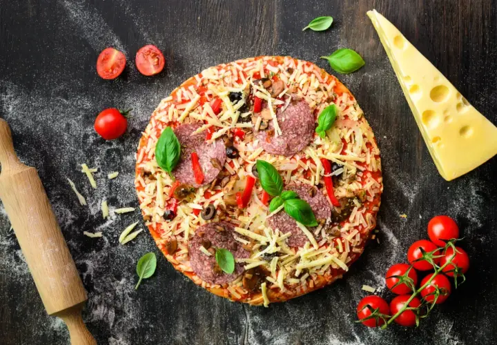 Gotowa pizza z salami, serem i warzywami, obok wałek, ser i pomidorki. Idealna do przygotowania z odpowiednią mąką do pizzy.
