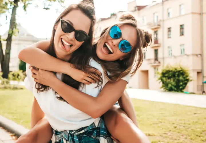 Dos amigas riendo y abrazándose, como si estuvieran escribiendo una carta a una amiga. Llevan gafas de sol y están al aire libre.
