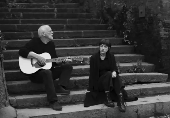 David Gilmour gra na gitarze, a obok niego siedzi jego córka.