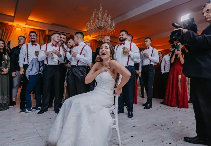 Radosna panna młoda na krześle, otoczona przez drużbów w czerwonych szelkach. Zastanawiasz się, ile kosztuje wesele na 50 osób?
