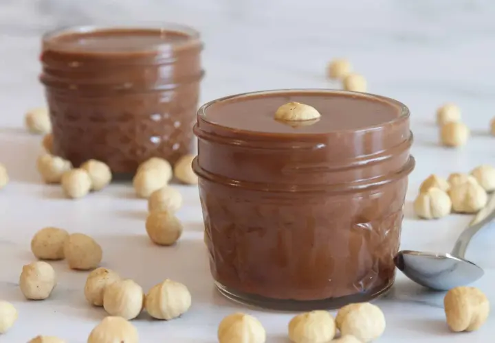 Dos tarros de deliciosa nutella vegana rodeados de avellanas. Una cucharilla plateada descansa cerca.