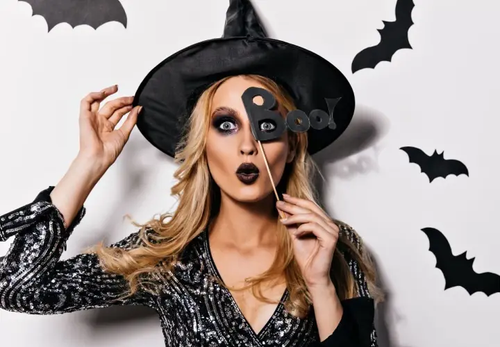 Kobieta w kapeluszu czarownicy z literami "Boo!" przed twarzą. Jej prosty makijaż czarownicy podkreśla oczy i usta.