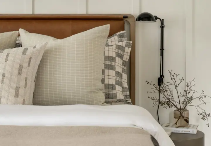 Studio McGee bed styling with layered pillows featuring subtle patterns and a plaid accent. A bedside lamp and delicate branches in a vase complete the serene look.