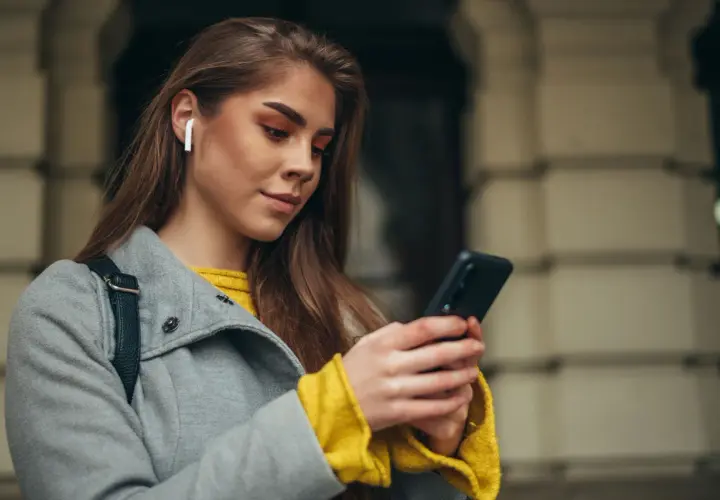 Kobieta z białymi słuchawkami bezprzewodowymi w uchu korzysta z telefonu Samsung. Dowiedz się, jak sparować słuchawki bezprzewodowe z telefonem Samsung.