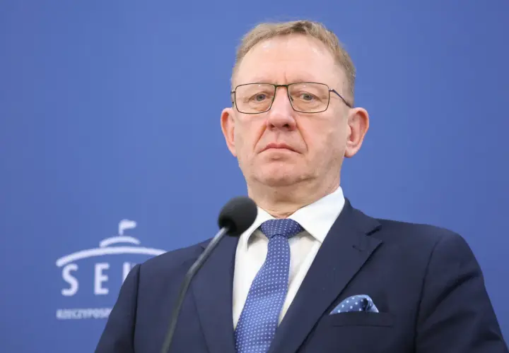 Mężczyzna w okularach i granatowym garniturze przed mikrofonem na tle niebieskiej ściany z logo Senatu.