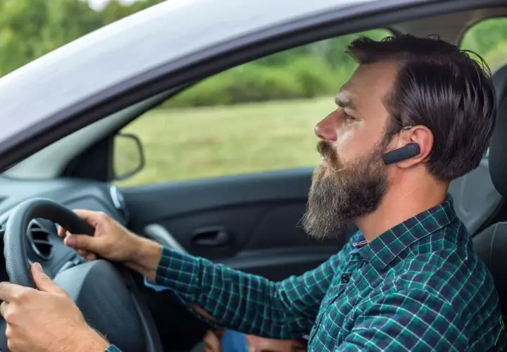 Mężczyzna z brodą i słuchawką Bluetooth w uchu prowadzi samochód. Dowiedz się, jak sparować telefon z samochodem, by bezpiecznie rozmawiać.