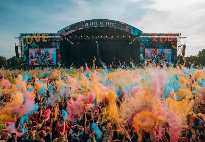 Une foule en liesse lance de la poudre colorée pour créer un festival de musique inoubliable. L'ambiance est électrique sous la scène "IN LOVE WE TRUST".