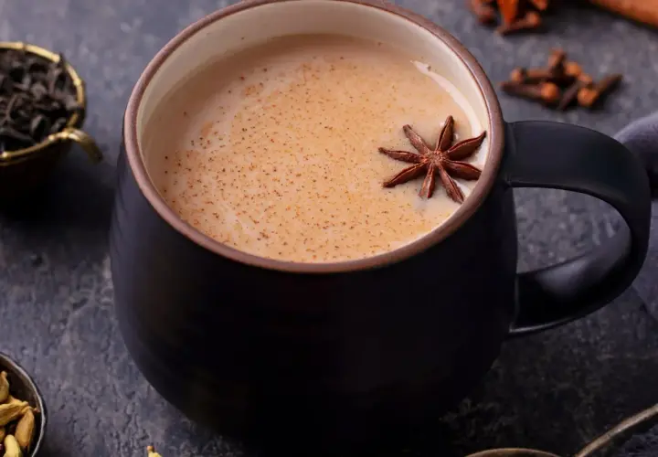 Aromaticzny napój, idealny na chłodne dni. Gorący **masala chai** w czarnej filiżance, ozdobiony anyżem. Obok liście herbaty, kardamon i goździki.