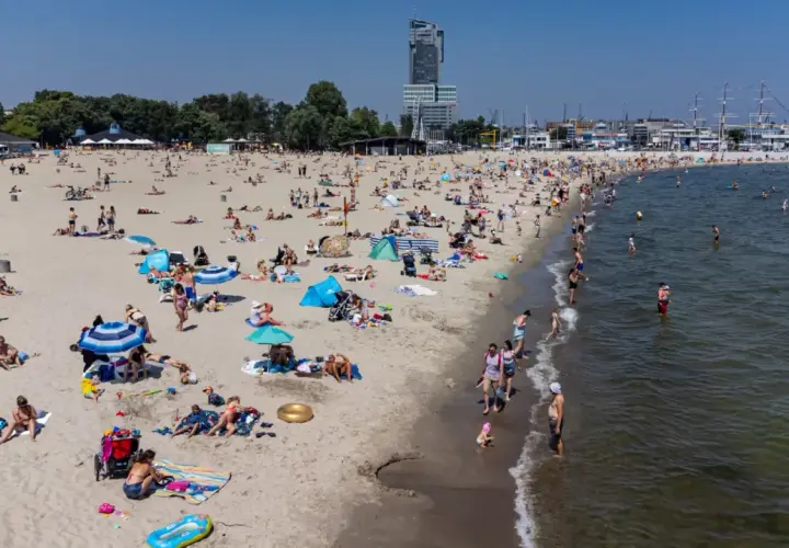 Słoneczna plaża w jednej z miejscowości koło Gdyni, pełna wypoczywających ludzi. W tle widać drapacz chmur i żaglowce.