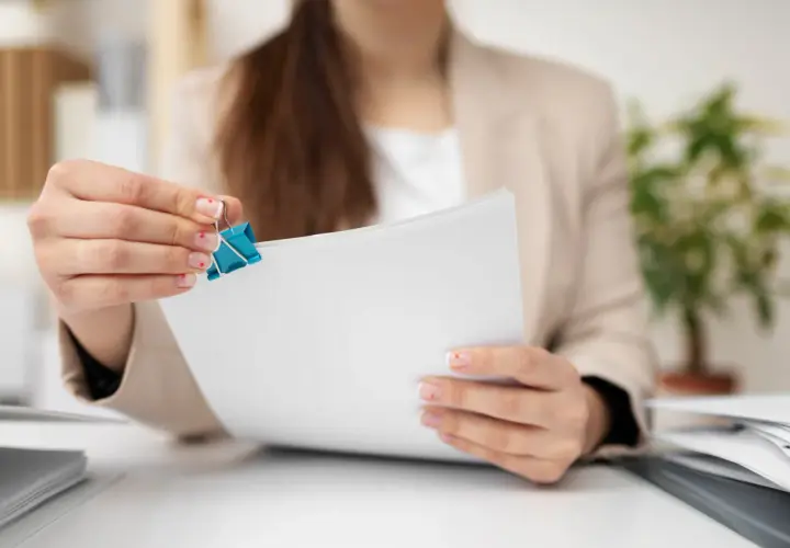 Kobieta w jasnym żakiecie trzyma niebieski klips do papieru na białych dokumentach.
