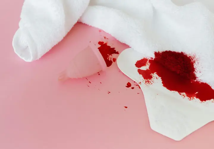 Sennik krew menstruacyjna: kubeczek i podpaska z plamami krwi na różowym tle.