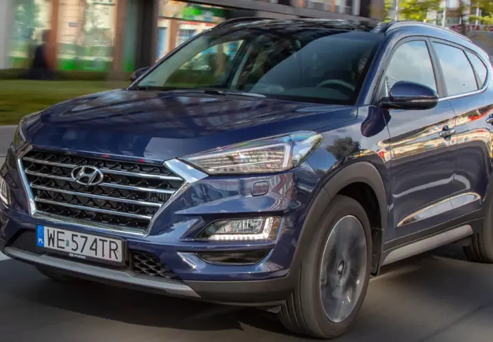 Nowy Hyundai Tucson w ciemnoniebieskim kolorze jedzie ulicą. Elegancka krata wlotu powietrza i nowoczesne reflektory podkreślają jego dynamiczny wygląd.
