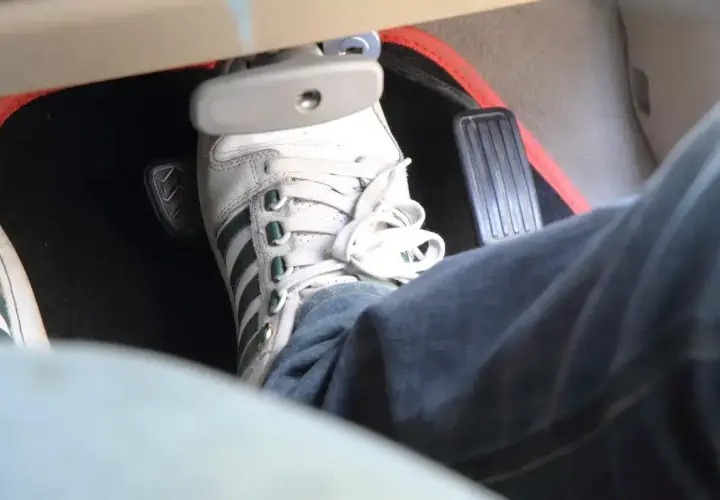 Un pied en basket Adidas appuie sur la pédale de frein, qui semble un peu molle. Le tapis de sol est noir avec un contour rouge.