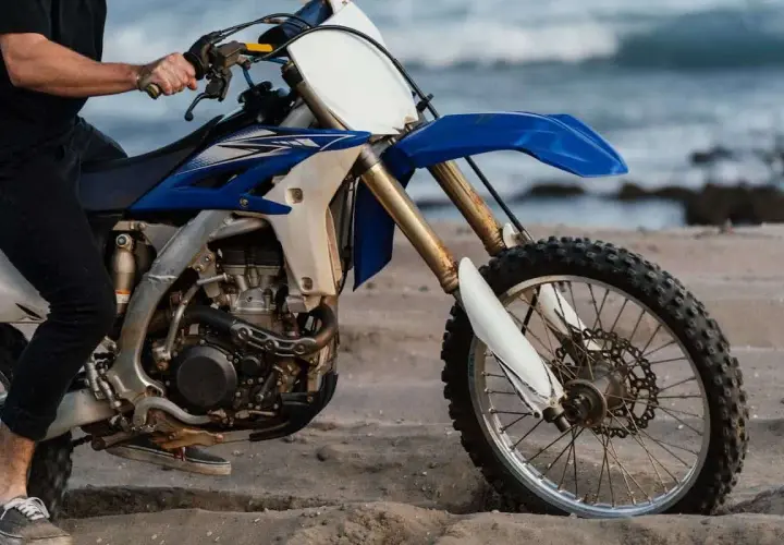 Persona sentada en una moto de cross azul y blanca en la arena, con el mar al fondo. ¿Cuánto paga un desguace por una moto?