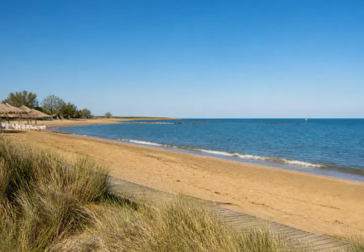 Plage de sable doré à Caorle, avec des parasols de chaume, des chaises blanches et la mer bleue sous un ciel clair.