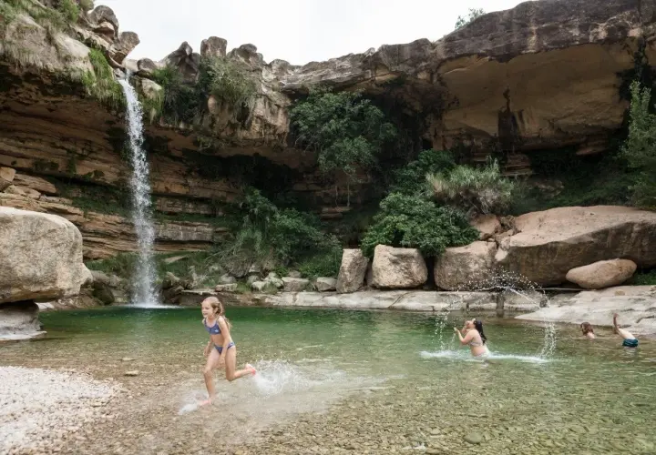 Niños disfrutan en una **piscina natural** bajo una cascada, rodeada de rocas y vegetación.