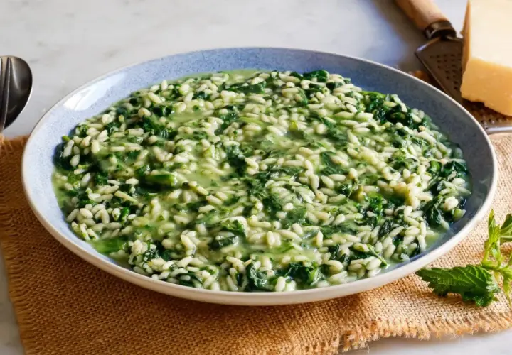 Un cremoso risotto con ortiche, servito in un piatto blu, con forchetta e coltello a lato. Un pezzo di parmigiano e una grattugia completano la scena.